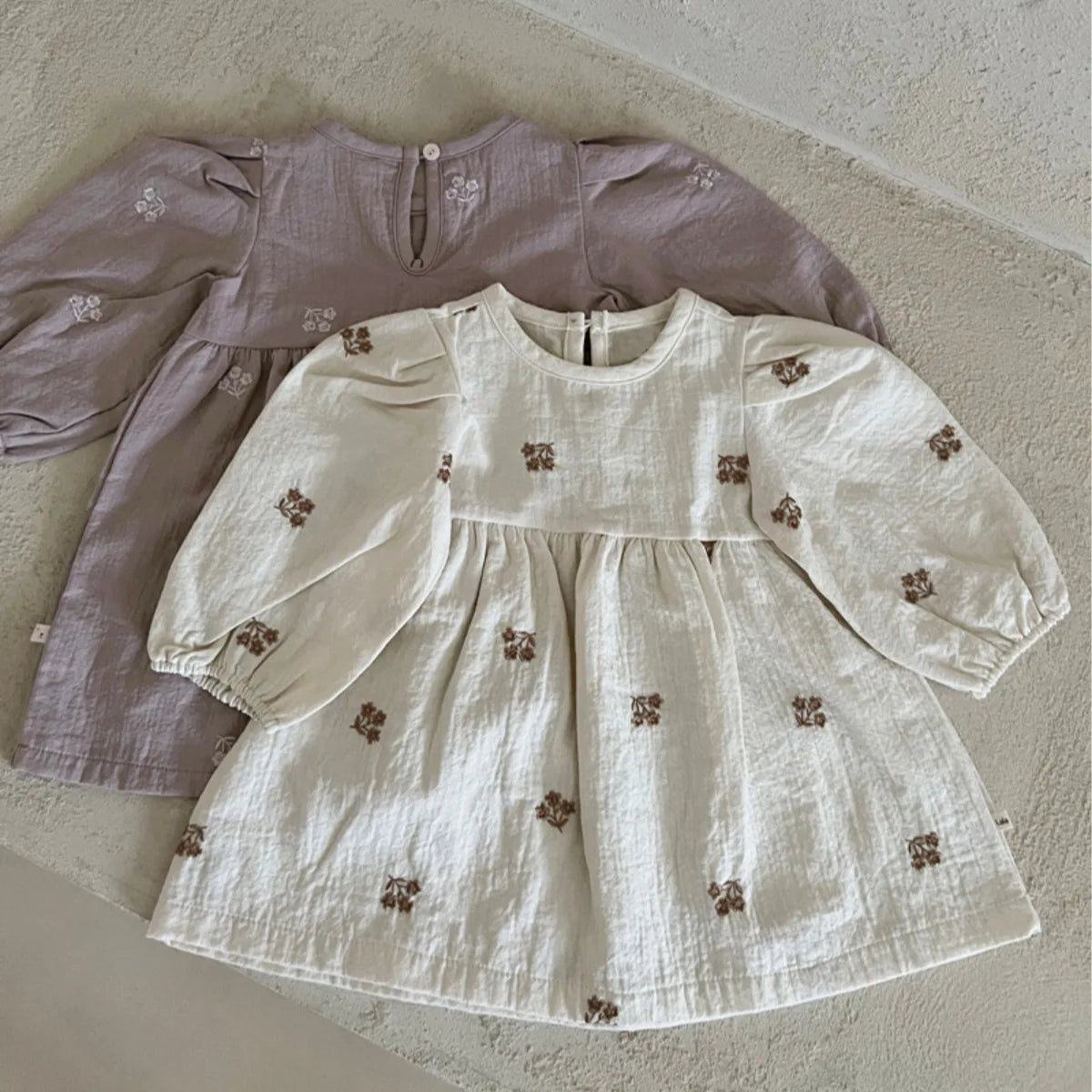 Two long-sleeve baby dresses laid flat on a textured beige surface. The front dress is off-white with small brown embroidered motifs, and the dress behind it is dusty mauve with similar embroidered details.