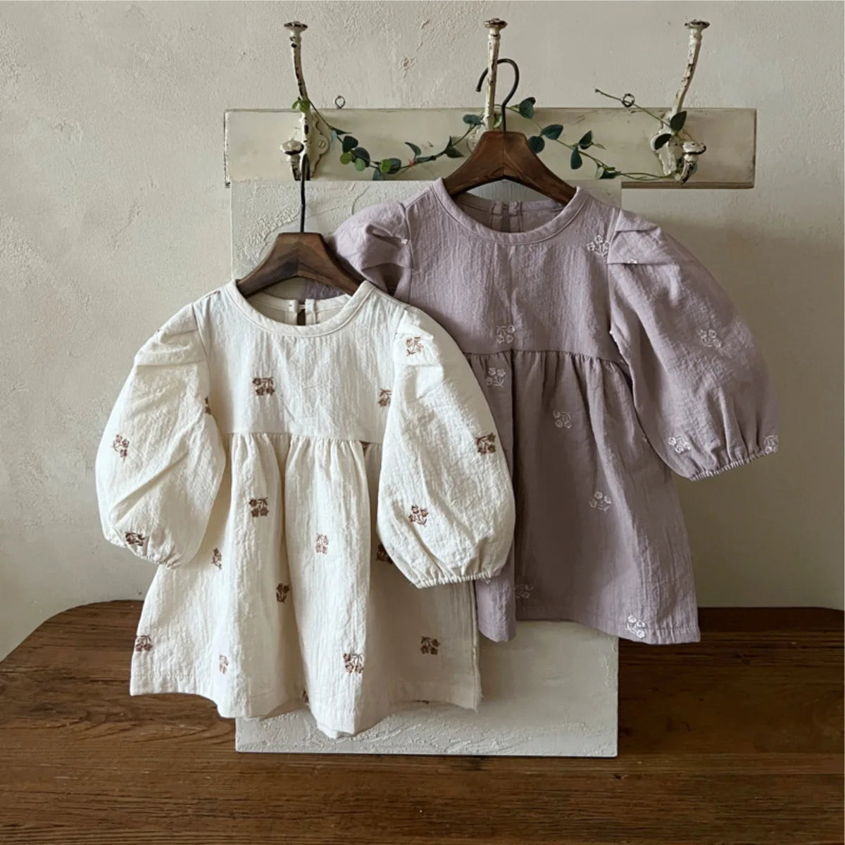 Two long-sleeve baby dresses hanging on wooden hangers on a wall rack. The front dress is off-white with small brown embroidered motifs, and the dress behind it is a dusty lavender with matching small embroidered motifs.